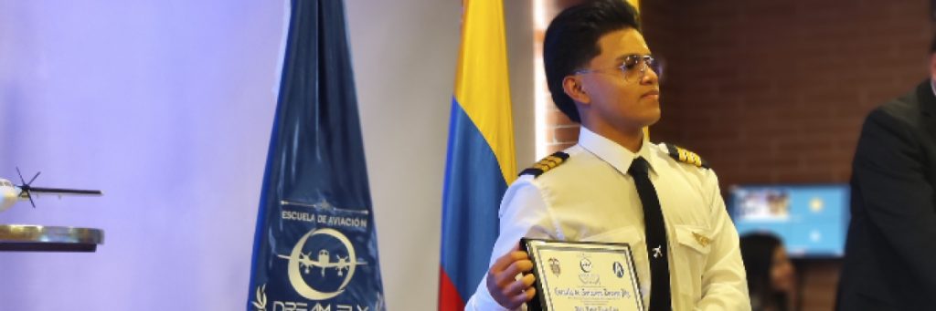 Estudiante piloto de aviación uniformado sosteniendo su certificado, con banderas de fondo, en la sección sobre beneficios de la certificación SMS para tu perfil profesional.