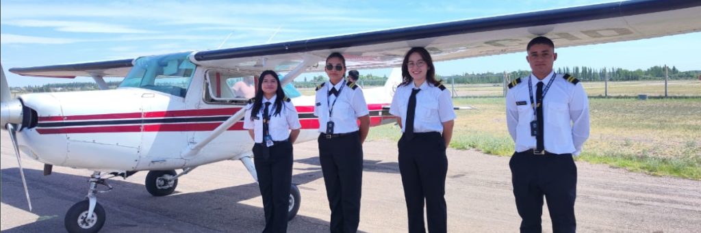 Cuatro estudiantes de aviación dos mujeres y dos hombres parados en formación frente a un aeronave en la pista como parte de su preparación profesional.