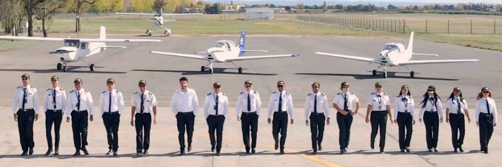 Grupo de aspirantes a piloto y estudiantes de aviación caminando en la pista de entrenamiento frente a aeronaves.