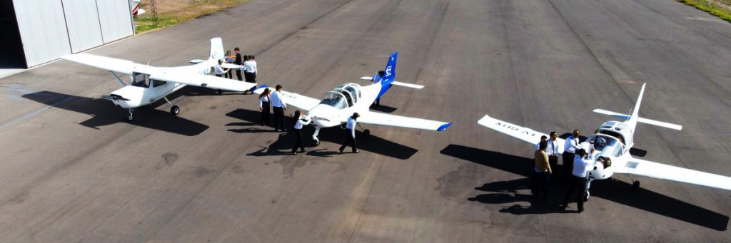 Grupo de estudiantes de aviación uniformados junto a tres de nuestras aeronaves en la pista de entrenamiento, representando la formación de excelencia y el inicio de la carrera profesional en Dream Fly.